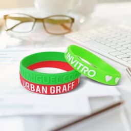 Three silicone wristbands in green, red, and white, featuring embossed text and a logo, resting on a desk with glasses and stationery.