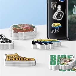A collection of custom shape die-cut stickers in various designs and colours, stacked near a smartphone with stickers on it.