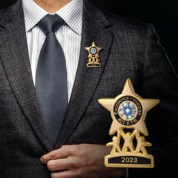 A brass soft enamel lapel pin in the shape of a star, featuring blue accents and a logo, worn on a man's suit jacket.