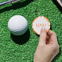 A hand holding an orange and white poker chip ball marker next to a white golf ball on artificial grass.