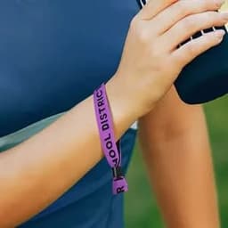 A person wearing a purple woven wristband on their wrist while holding a drink bottle. The wristband has a logo.