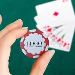 A hand holding a red and grey custom plastic poker chip with a logo, set against a backdrop of playing cards.