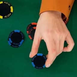 A hand placing a blue poker chip on a green table, surrounded by red and black chips. The chips have a decorative border.