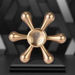 A bronze fidget spinner in a playful rudder shape, displayed on a black pedestal. Features a logo at the centre.