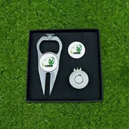 A silver golf tee marker, a ball position marker, and a clip presented in a black box on grass.
