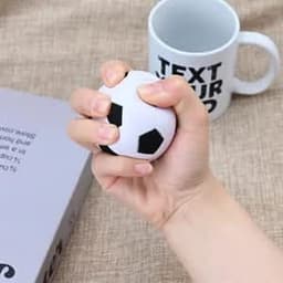 A hand squeezing a white soccer stress ball with black pentagon patterns, next to a mug.