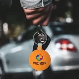 A hand holding a round, orange silicone key tag attached to a car key, with a logo present on the tag.
