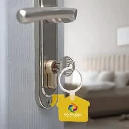 A house-shaped silicone keychain in yellow, attached to a key, inserted in a door lock. The background features a blurred room.
