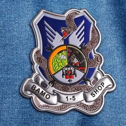 A metallic badge featuring a blue shield, intricate designs, and a logo. It has a snake and banner at the base.