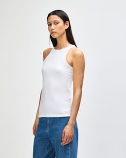 A woman in a white racerneck tank top stands sideways, showcasing the fit. The top has a subtle design and a brand logo.