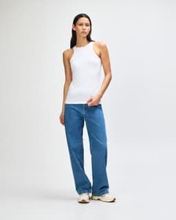A white women's racerneck tank top worn with light blue wide-leg jeans, featuring a logo on the tank.