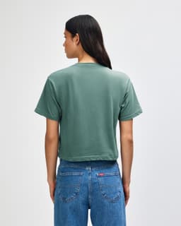 A green women's boxy t-shirt, viewed from the back, paired with light-coloured jeans. It features a branding logo.