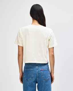 A beige women's boxy t-shirt with short sleeves, viewed from the back, featuring a logo. Casual style, perfect for layering.