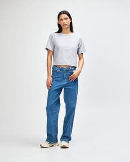 A woman wearing a grey boxy t-shirt and blue jeans, standing against a plain background. The shirt has a logo.