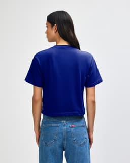 A blue boxy t-shirt with short sleeves and a back view. The model wears high-waisted jeans and has curly hair.