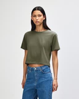 A woman wears an olive green boxy t-shirt, styled with light blue jeans, against a plain background.