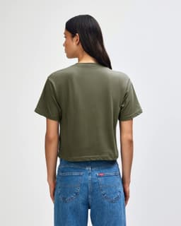 A back view of a women's olive green boxy T-shirt, paired with light blue cargo jeans. Features a logo.