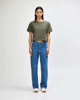 A woman stands wearing an olive green boxy t-shirt and light blue cargo jeans, paired with black and white sneakers.
