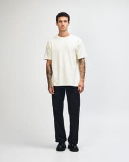 A unisex heavyweight cotton T-shirt in cream, paired with blue cargo pants and sneakers. The model has curly hair and a neutral pose.