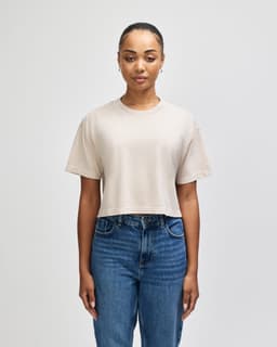 A cropped boxy t-shirt in a neutral beige colour, featured on a model wearing blue jeans. The shirt has a logo.