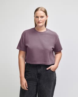 A women's cropped boxy T-shirt in a muted purple shade, featuring a logo. The model is styled with jeans.
