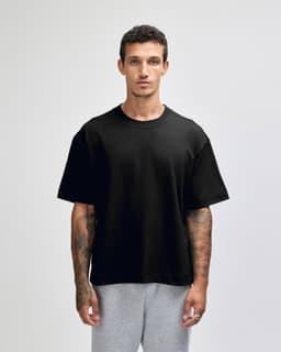 A black unisex heavyweight cotton boxy t-shirt displayed on a model with tattoos, paired with grey trousers.