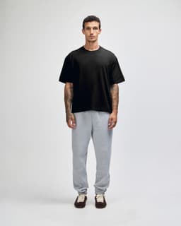 A man stands wearing a black heavyweight cotton unisex boxy t-shirt and grey sweatpants. The image features a logo on the t-shirt.