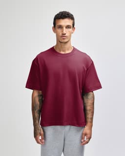 A maroon heavyweight cotton unisex boxy t-shirt displayed on a model with tattoos, paired with grey pants.