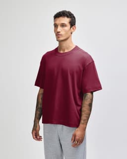 A maroon unisex boxy T-shirt displayed on a model, featuring a branding logo, paired with grey pants.