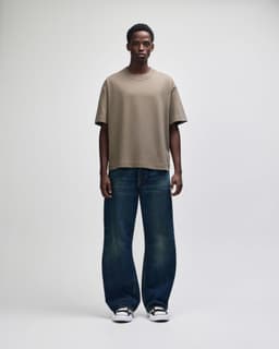 A unisex boxy T-shirt in a shade of brown, worn with loose-fitting blue jeans. The model stands upright against a plain background.