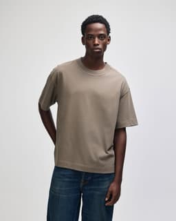 A unisex boxy T-shirt in a muted olive green colour, worn by a model standing against a neutral background.