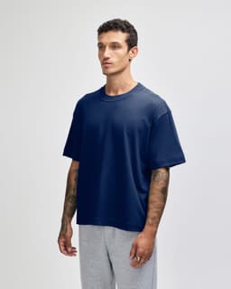 A navy unisex boxy T-shirt made of heavyweight cotton, featuring a logo. Worn with grey trousers.