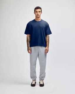 A man stands wearing a navy blue heavyweight cotton unisex boxy t-shirt and grey sweatpants. The shirt features a logo.