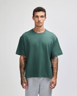 A green heavyweight cotton unisex boxy T-shirt displayed on a model with tattoos, paired with grey pants.