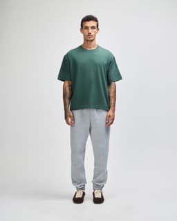 A unisex boxy t-shirt in green, paired with grey sweatpants, worn by a model standing against a plain background.
