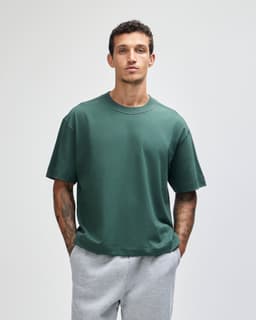 A green heavyweight cotton unisex boxy t-shirt, worn by a model with short hair, paired with grey sweatpants.