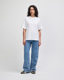 A woman stands wearing a white, heavyweight cotton, unisex boxy t-shirt paired with blue wide-leg jeans.
