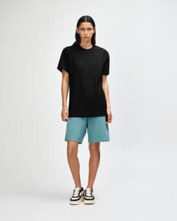 A person wearing a black unisex pique mockneck t-shirt paired with teal shorts and black sneakers, standing against a neutral background.