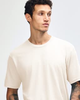 A model wearing an unbranded beige unisex short sleeve t-shirt and loose blue jeans, standing upright.