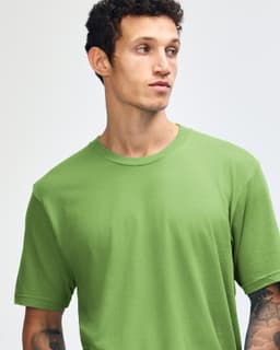 A model stands wearing a green unisex fine jersey short sleeve T-shirt and loose-fitting blue jeans.