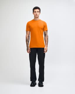 An orange unisex short sleeve t-shirt worn by a model, paired with beige cargo pants. The t-shirt features a logo.