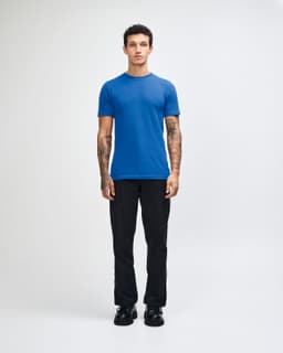 A blue short sleeve t-shirt displayed on a model wearing black pants, featuring a logo on the t-shirt.