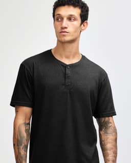 A black unisex Henley T-shirt featuring short sleeves and a button-up neck. The model has tattoos on their arms.