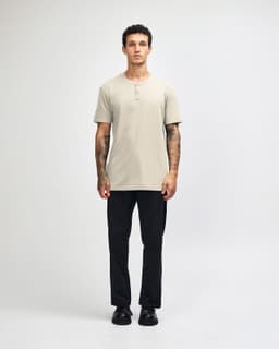 A beige unisex Henley T-shirt featured on a model paired with light blue jeans and black sneakers. The shirt has a logo.