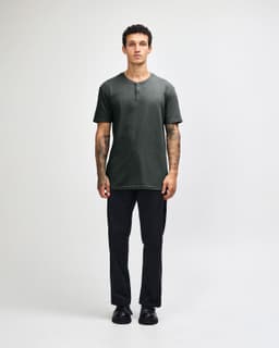A man stands wearing a dark grey unisex Henley T-shirt paired with light blue jeans and black sneakers.