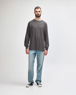 A long-sleeve T-shirt in grey worn by a model, paired with black jeans and sneakers. The shirt features a logo.