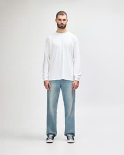 A white long sleeve T-shirt worn by a man, paired with black pants and shoes, standing against a light background.