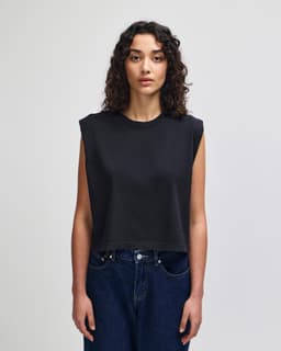 A women's muscle t-shirt in dark grey, featuring a cropped, sleeveless design. The model has curly hair and wears jeans.