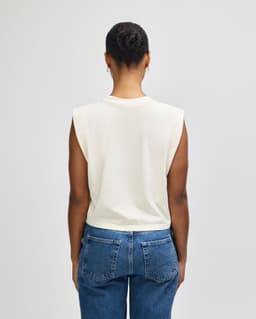 A women's muscle T-shirt in cream, featuring a sleeveless design and a relaxed fit. The shirt has a visible logo.