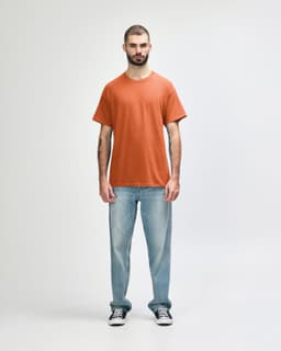 An adult wearing an orange Gildan Heavy Cotton T-shirt, paired with black pants and black shoes. The shirt has a logo.